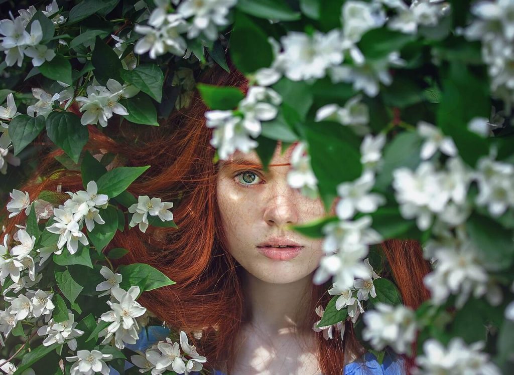 a beautiful girl with red hair looking through a beautiful flower bush - this picture is used in article about omega 3 beauty benefits