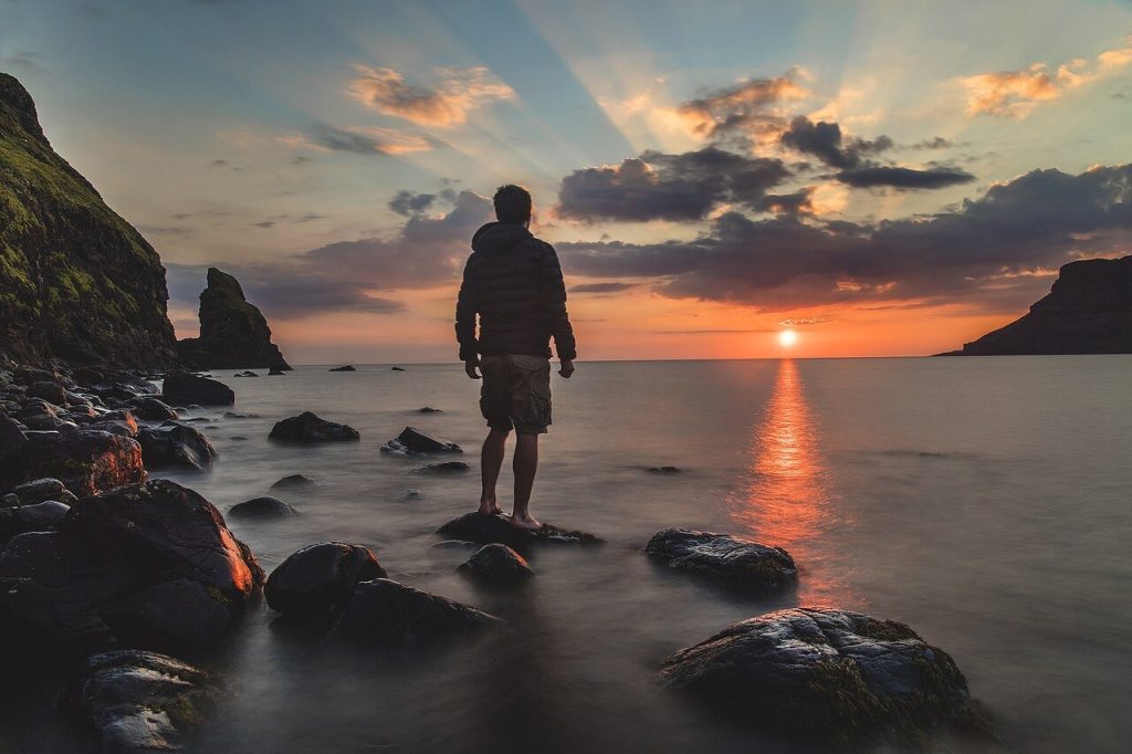 a young man standing on a rock on the sea side watching the sunset in the sky - this picture is used in article about the benefits of omega 3 for men