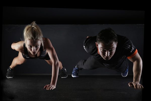 a blonde woman and a dark haired man doing one arm push ups in a dark background - the picture is used in an article about the benefits of omega 3 in fitness