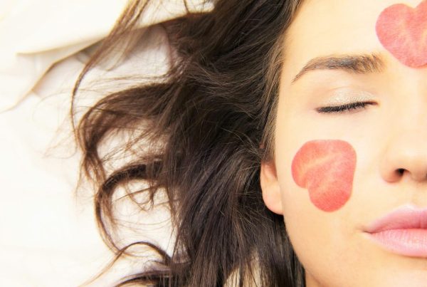 woman laying and only her face is seen with closed eyes and rose petals on her cheek and forehead - this piture is used in article about omega 3 benefits for rosacea