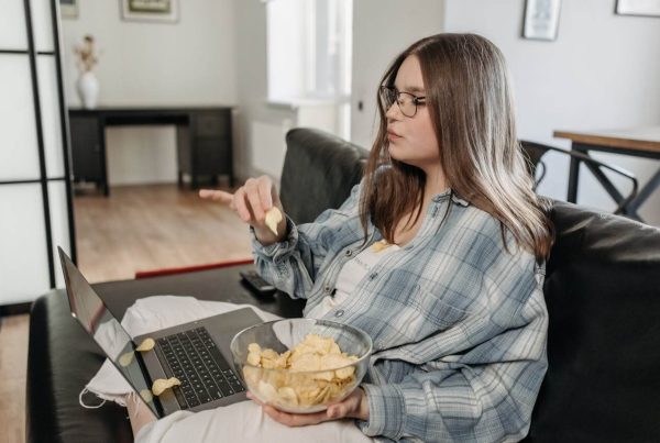 Woman Holding a Bowl of Potato Chips Sitting on a Couch - used in article on obesity and overweight overview